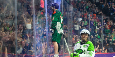 Halifax Thunderbird Jake Withers celebrates a goal by standing on a ledge on the boards as Saskatchewan Rush captain Ryan Keenan looks on.